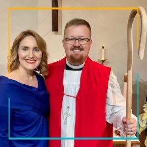 Bishop Jonathan standing with his staff, red vestment, next to his wife, Ivey in a bright blue dress